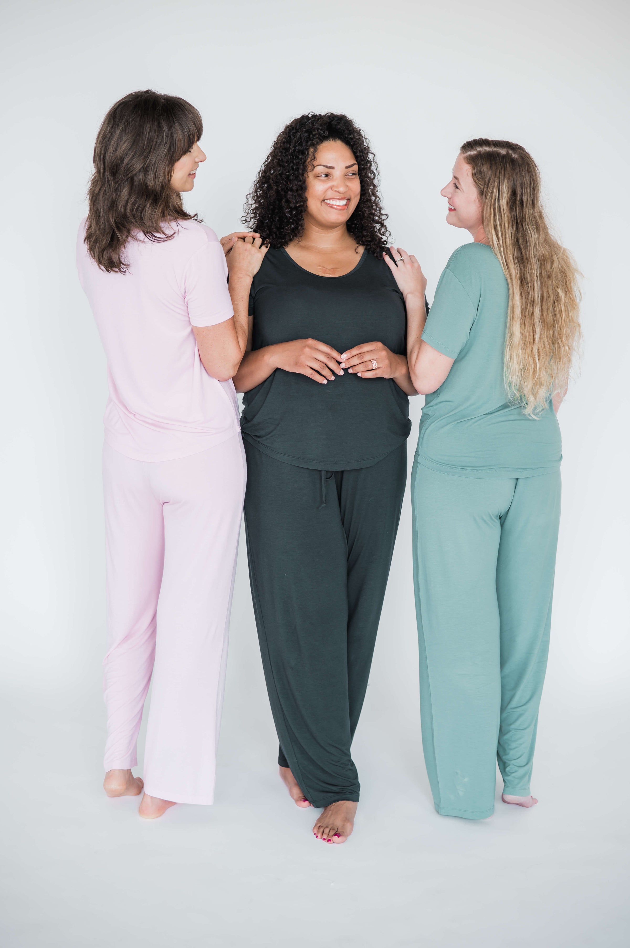 3 women stand touching hands in bamboo postpartum pajama sets. One in pink, one pregnant in black, one in sage green. Bamboo pajama sets designed for postpartum nursing, c-section recovery, and long nights with a newborn.