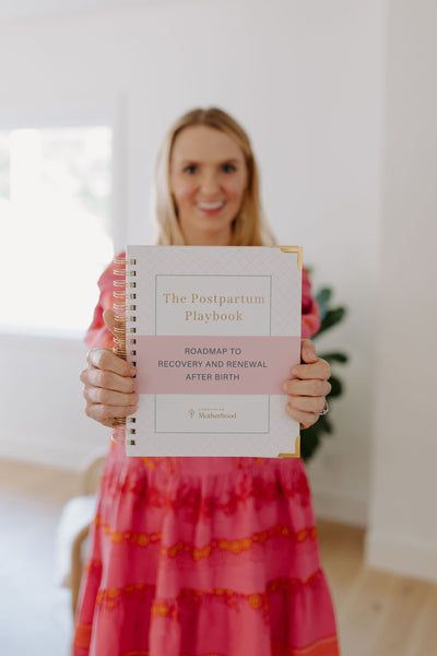 Woman holding a book titled 'The Postpartum Playbook' in a bright room.