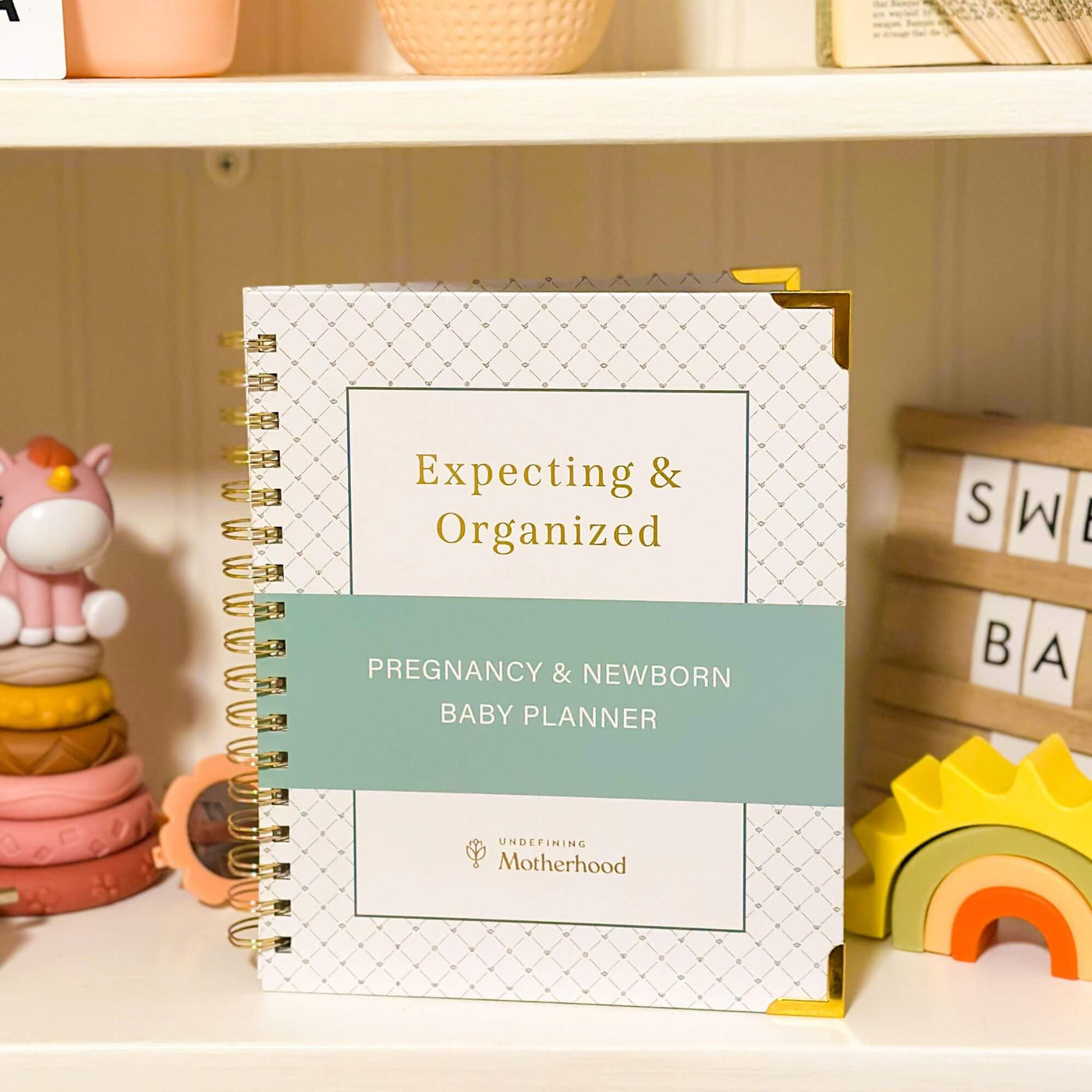 A white pregnancy planner with gold binding sits in the middle of a bookshelf, between baby toys and a letterboard that says sweet baby. The planner has a white cover with a green stripe and a small pattern design on the border. The cover of the book says Expecting & Organized: Pregnancy & Newborn Baby Planner by Undefining Motherhood. The title and company logo are gold foil stamped into the white cover, and the edges are secured with gold edging.