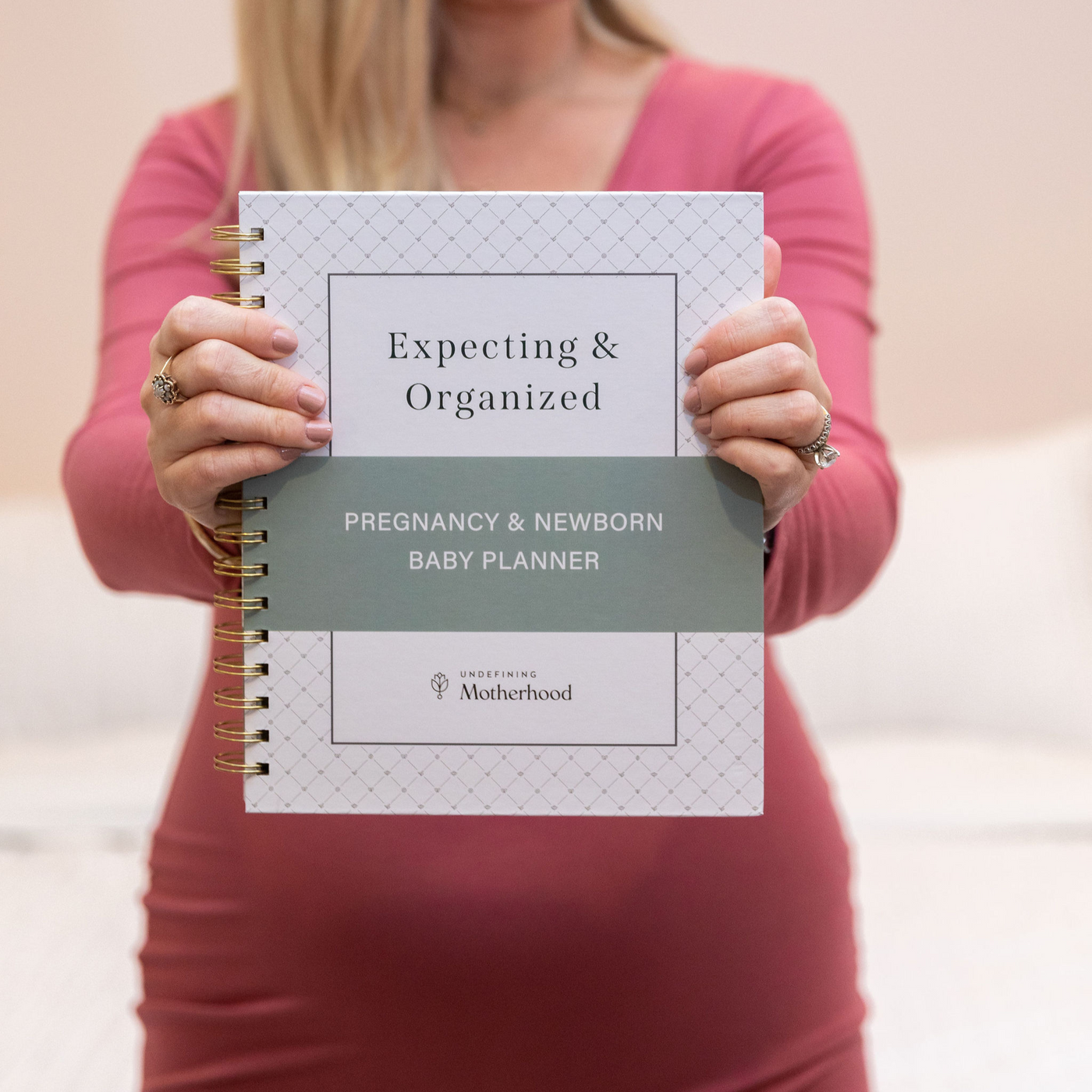 A woman's hands hold a white pregnancy planner in front of her small baby bump. The baby planner is white with a hardcover and gold o-ring binding, with a soft green stripe in the middle. It says Expecting & Organized: Pregnancy & Newborn Baby Planner by Undefining Motherhood